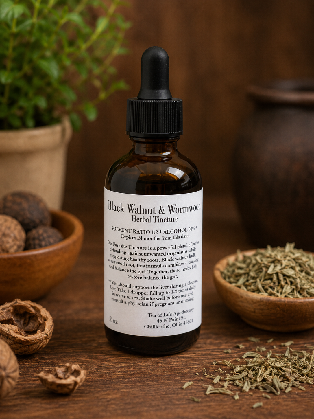 Tea of Life Apothecary Black Walnut and Wormwood Herbal Tincture in amber dropper bottle displayed on a rustic wooden table with dried wormwood herbs and black walnuts in a warm apothecary setting.