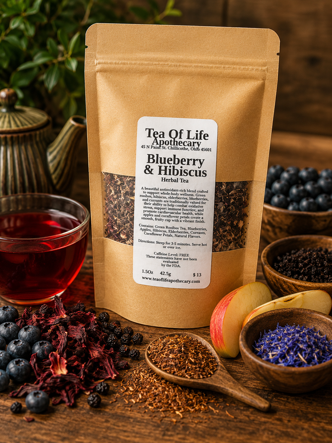 Tea of Life Apothecary Blueberry & Hibiscus Herbal Tea displayed with fresh blueberries, dried hibiscus flowers, green rooibos tea, dried elderberries, sliced apples, blue cornflower petals, and a vibrant ruby-red cup of herbal tea on a rustic wooden table. This naturally caffeine-free antioxidant tea blend offers a fruity, refreshing flavor that can be enjoyed hot or iced.