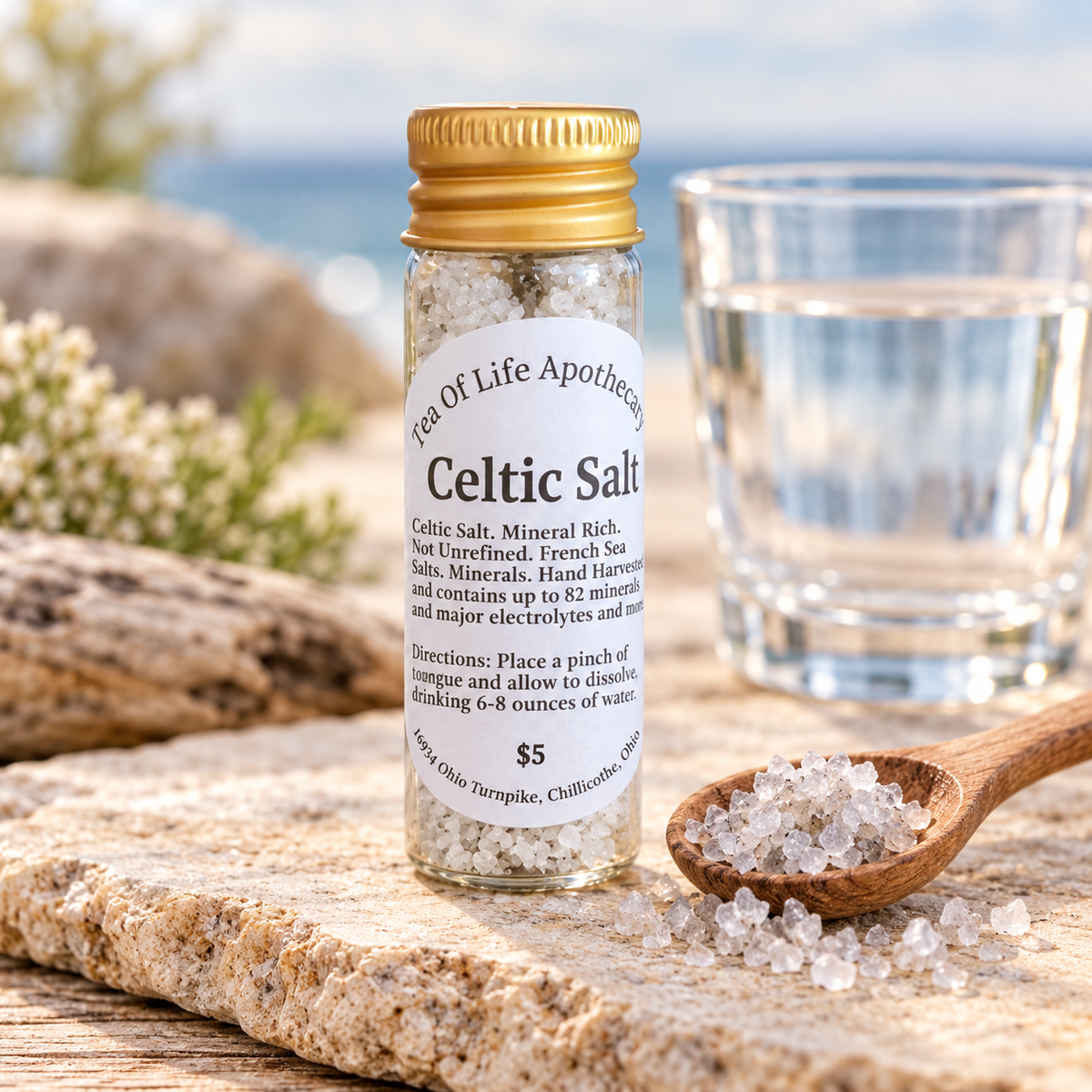 Tea of Life Apothecary Celtic Salt in a glass vial with coarse mineral-rich sea salt, a wooden measuring spoon, and a fresh glass of water.