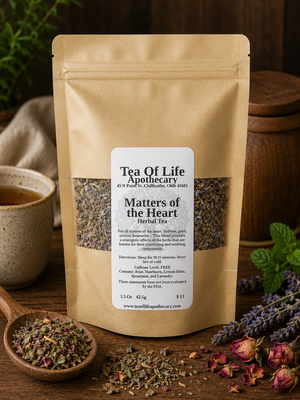A stand-up kraft paper pouch labeled 'Tea Of Life Apothecary Matters of the Heart Herbal Tea' stands on a wooden surface with loose tea, a cup, herbs, and flowers around it.