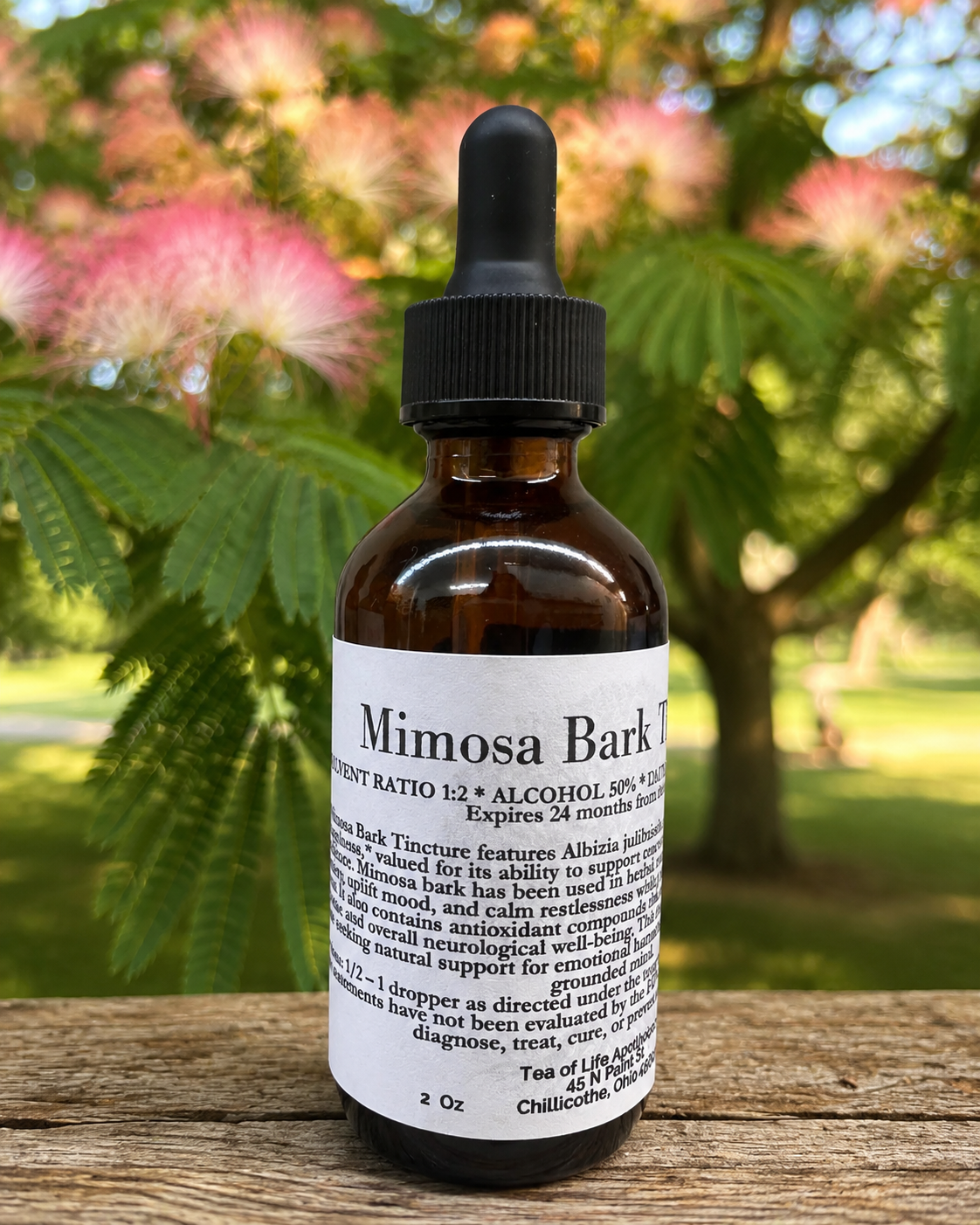 Tea of Life Apothecary Mimosa Bark Tincture in a 2 oz amber glass dropper bottle displayed outdoors with a softly blurred mimosa tree in bloom. Herbal tincture made with mimosa bark traditionally used to support emotional balance, relaxation, and nervous system wellness.