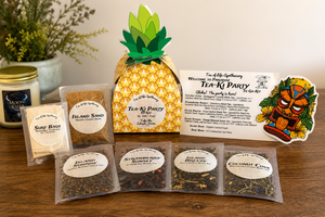 A tea gift set arranged on a wooden table, including a pineapple-shaped box, several tea bags, a candle jar, a small bag of sand, and printed cards and stickers. The visible product branding reads Tea of Life Apothecary.