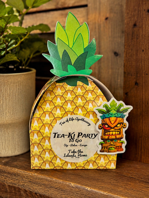A pineapple-shaped paper party favor box with a tiki character sticker, displayed on a wooden surface next to a potted plant.