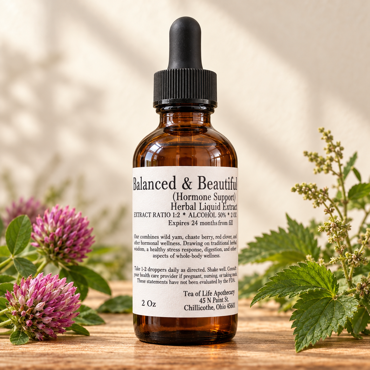 Balanced & Beautiful herbal tincture by Tea of Life Apothecary, pictured in a 2 oz amber dropper bottle with red clover flowers and green herbs on a wooden surface.