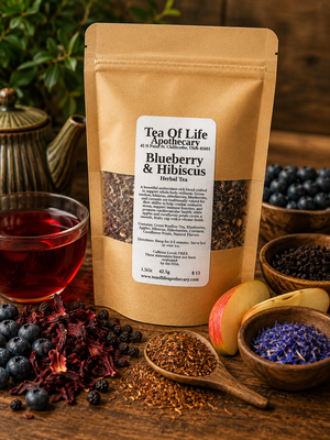 Tea of Life Apothecary Blueberry & Hibiscus Herbal Tea displayed with fresh blueberries, dried hibiscus flowers, green rooibos tea, dried elderberries, sliced apples, blue cornflower petals, and a vibrant ruby-red cup of herbal tea on a rustic wooden table. This naturally caffeine-free antioxidant tea blend offers a fruity, refreshing flavor that can be enjoyed hot or iced.