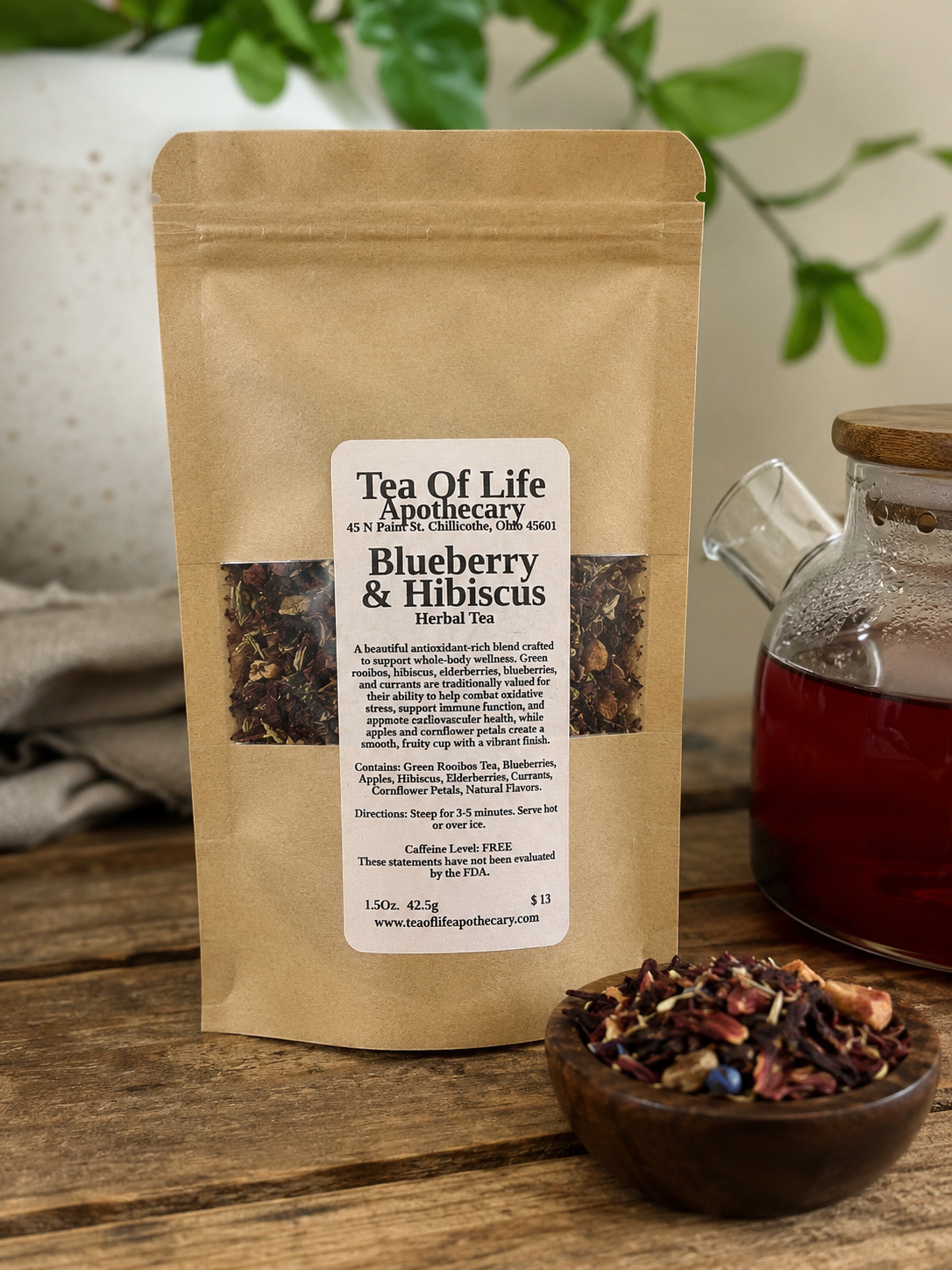Blueberry & Hibiscus Rooibos Herbal Tea by Tea of Life Apothecary displayed in a kraft paper tea pouch on a rustic wooden table beside a glass teapot and loose tea blend, featuring antioxidant-rich ingredients including blueberries, hibiscus, elderberries, currants, and green rooibos tea.