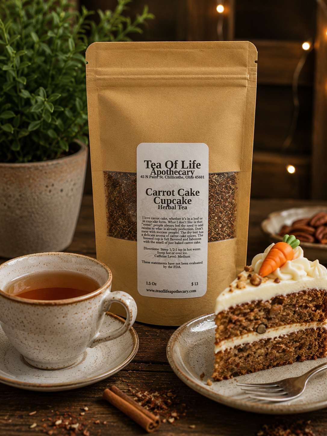 A stand-up kraft pouch of Tea Of Life Apothecary Carrot Cake Cupcake Herbal Tea stands on a wooden table beside a cup of tea, a slice of carrot cake on a plate, cinnamon sticks, and a potted plant. The pouch has a white label with black text and two clear windows showing the tea and loose botanicals inside.