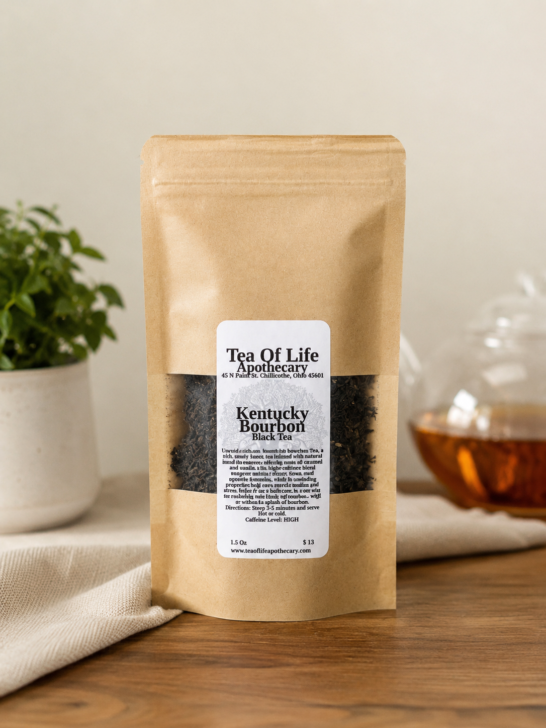 Kentucky Bourbon Black Tea by Tea of Life Apothecary in a kraft stand-up pouch, featuring rich black tea with notes of bourbon, caramel, and vanilla, displayed on a rustic wooden table beside a glass teapot and greenery.