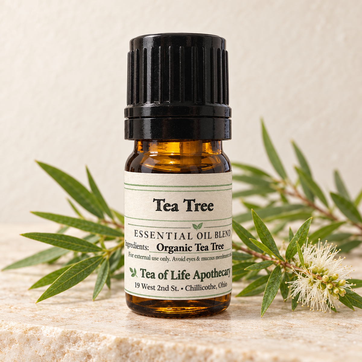 Tea of Life Apothecary Organic Tea Tree Essential Oil in a 5 mL amber glass bottle, displayed with authentic melaleuca leaves and white blossoms.