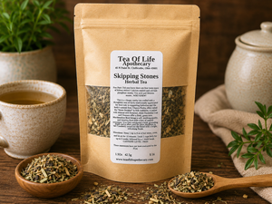 Tea of Life Apothecary Skipping Stones Herbal Tea displayed on a rustic wooden table with loose herbal tea, a steaming cup of tea, and natural botanical accents. This caffeine-free herbal blend features traditional herbs including chanca piedra, horsetail, dandelion leaf, cleavers, marshmallow root, uva ursi, licorice root, hibiscus, and cranberry in a warm apothecary-inspired setting.